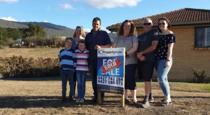 a family of 7 standing next to a PropertyNow for sale sign with a sticker that says 'SOLD' on it