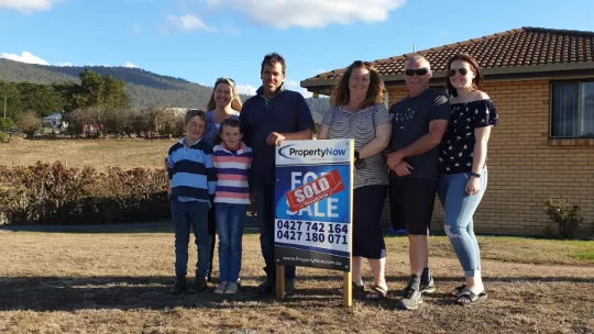 a family of 7 standing next to a PropertyNow for sale sign with a sticker that says 'SOLD' on it