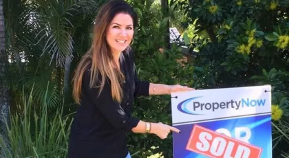 a woman pointing to a PropertyNow for sale sign with the word 'SOLD' added to it