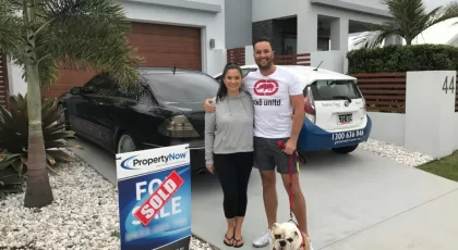 a woman and a man with their dog standing next to a PropertyNow for sale sign with the word 'SOLD' added to it