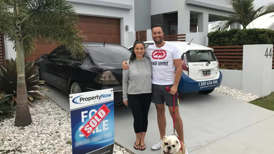 a woman and a man with their dog standing next to a PropertyNow for sale sign with the word 'SOLD' added to it